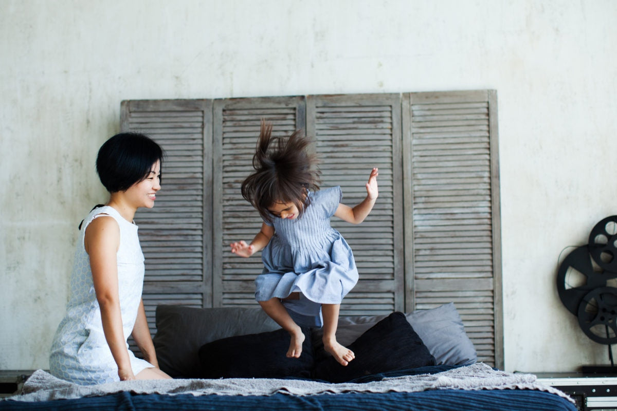 Happy little child girl jumping on bed and mother looking at her happily