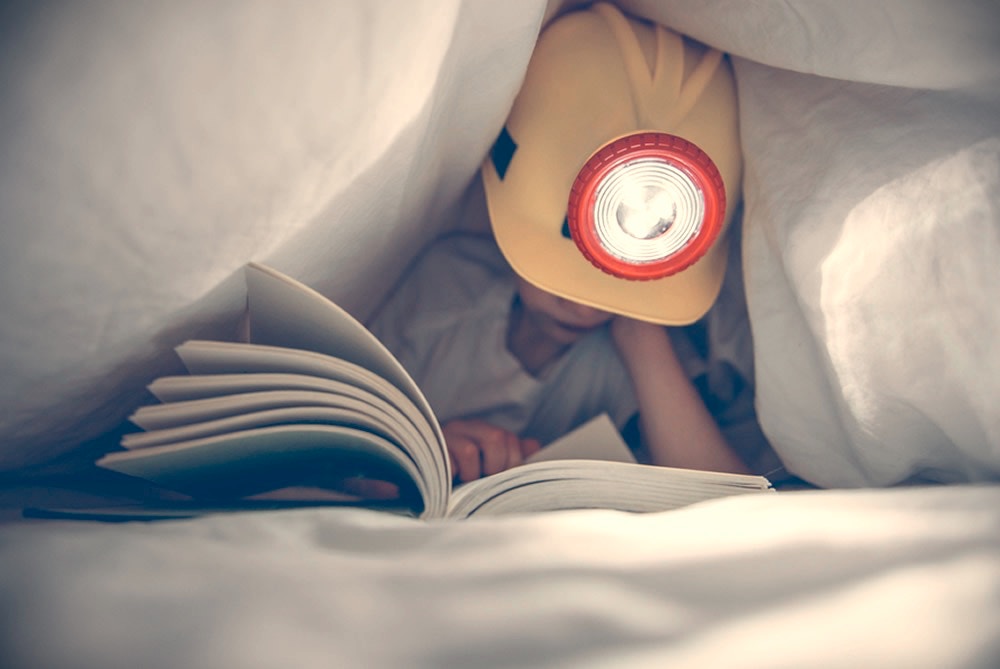 young boy with construction hat under his bed sheets reading a book with a flash light