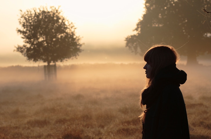 Young Woman Walking On A Road Through A Forest Filled With Fog And Sunlight