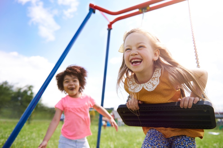 Two happy smiling little toddlers girls friends swinging on swings at playground outside