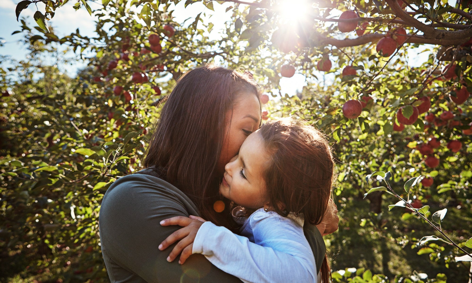 mother is kissing her daughter on the cheek, apple trees on the background