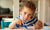 young boy drawing with crayons on sheets of white paper on a table